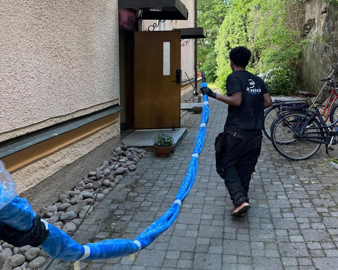 En arbetare i svart t-shirt bär en blå slang mot en husingång.